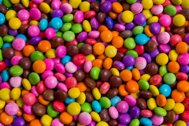 Assorted brightly colored gummy sweets including rings, fruit slices, and worms piled together in a vibrant display. The sweets are covered in sugar and arranged in a market-style setting with a cheerful and playful atmosphere. No visible text.