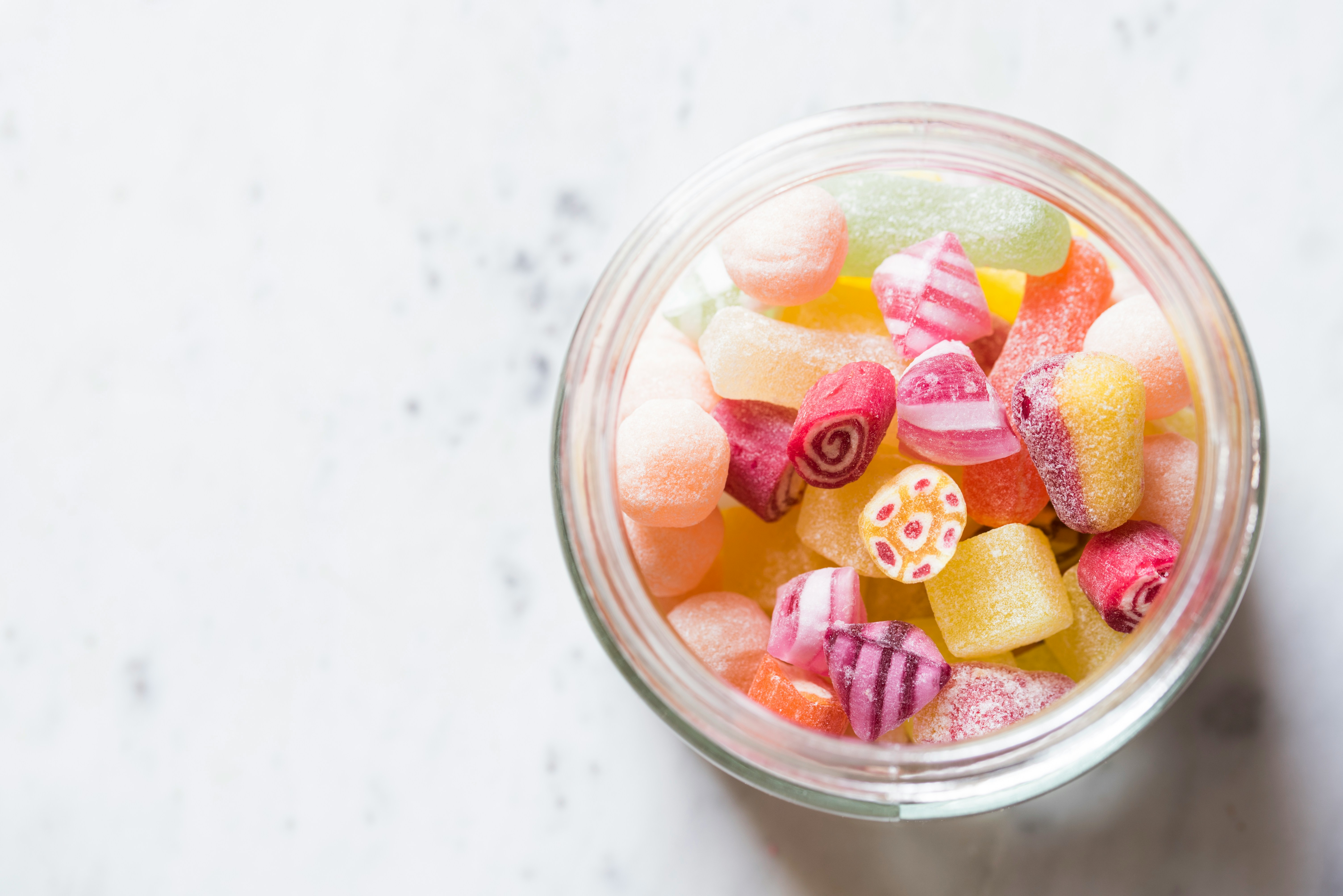 Glass jar filled with colorful assorted sweets sitting on a bright table surrounded by scattered candies. The scene feels cheerful and inviting, evoking a sense of nostalgia and delight. No visible text.