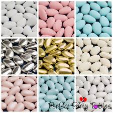 Sugared almonds arranged in elegant white and pastel packaging on a wedding table adorned with fresh flowers and satin ribbons. The setting is festive and romantic, creating a joyful and celebratory mood. No visible text.